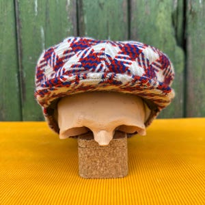May include: A newsboy cap with a red, white, and blue plaid pattern. The cap is displayed on a mannequin head, set against a yellow surface and a green wooden background. The cap's design is a classic style.