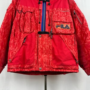 Puede incluir: Chaqueta de esqu&iacute; FILA roja con un patr&oacute;n geom&eacute;trico. La chaqueta tiene m&uacute;ltiples bolsillos, una cremallera azul y el logotipo de FILA en el pecho. Dise&ntilde;ada para el fr&iacute;o.