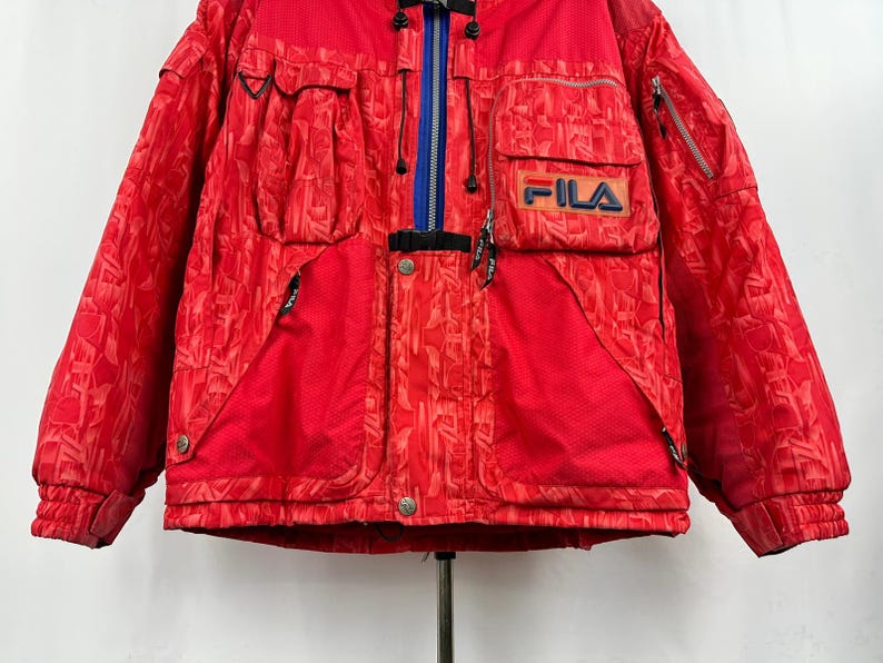 Puede incluir: Chaqueta de esqu&iacute; FILA roja con un patr&oacute;n geom&eacute;trico. La chaqueta tiene m&uacute;ltiples bolsillos, una cremallera azul y el logotipo de FILA en el pecho. Dise&ntilde;ada para el fr&iacute;o.