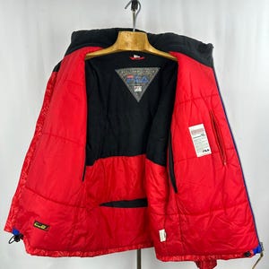 Puede incluir: Chaqueta de invierno FILA roja y negra con capucha negra y cremalleras azules. La chaqueta est&aacute; abierta, revelando un forro interior negro y un logotipo triangular. La chaqueta est&aacute; colgada de una percha de madera.