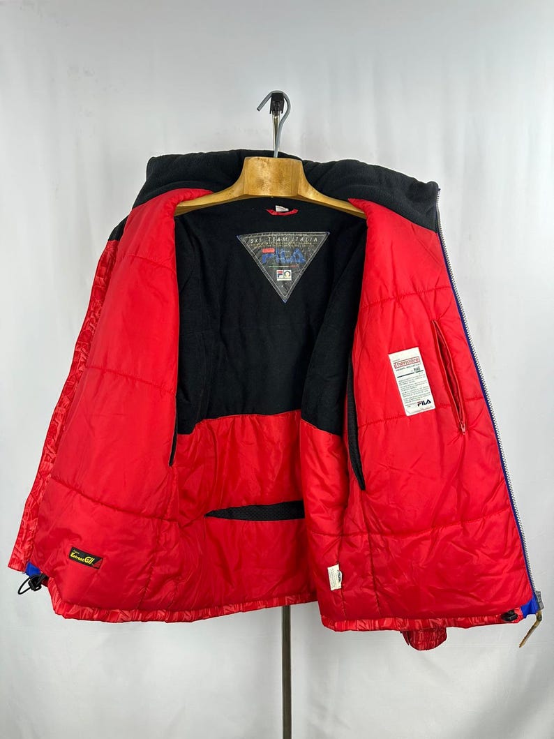 Puede incluir: Chaqueta de invierno FILA roja y negra con capucha negra y cremalleras azules. La chaqueta est&aacute; abierta, revelando un forro interior negro y un logotipo triangular. La chaqueta est&aacute; colgada de una percha de madera.