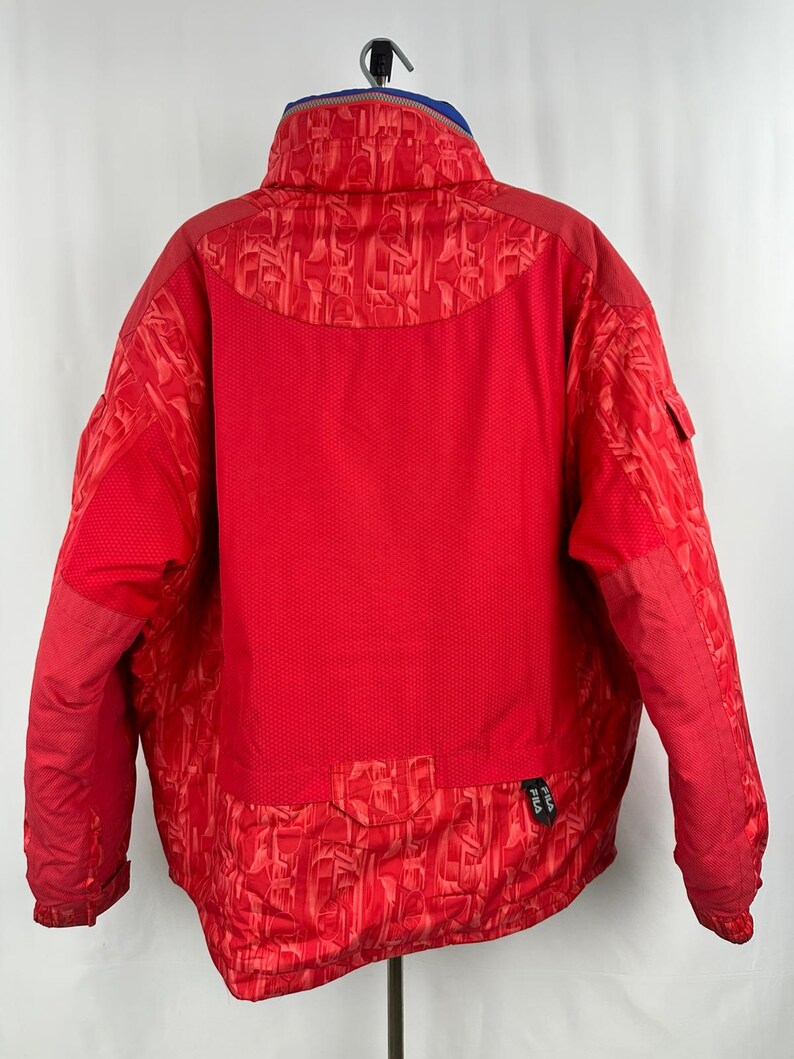 Puede incluir: Chaqueta de invierno roja brillante con cuello alto y forro interior azul. La chaqueta presenta un patr&oacute;n texturizado en los hombros y la parte inferior de la espalda, con un panel de malla en la parte superior de la espalda. El logotipo de Fila es visible en la parte inferior derecha.