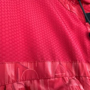 Puede incluir: Primer plano de una tela roja con un patr&oacute;n de panal. La tela tiene una superficie texturizada y forma parte de una prenda. Un cord&oacute;n negro y una pieza de pl&aacute;stico negro son visibles a la derecha.