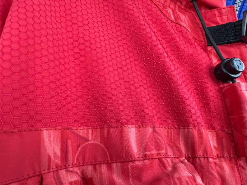 Puede incluir: Primer plano de una tela roja con un patr&oacute;n de panal. La tela tiene una superficie texturizada y forma parte de una prenda. Un cord&oacute;n negro y una pieza de pl&aacute;stico negro son visibles a la derecha.