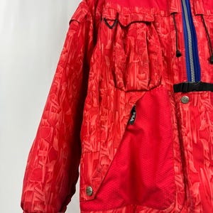 Puede incluir: Chaqueta de esqu&iacute; Fila roja con un patr&oacute;n geom&eacute;trico. La chaqueta presenta una cremallera azul, detalles en negro y m&uacute;ltiples bolsillos. Tiene un cuello alto y cierre de bot&oacute;n.