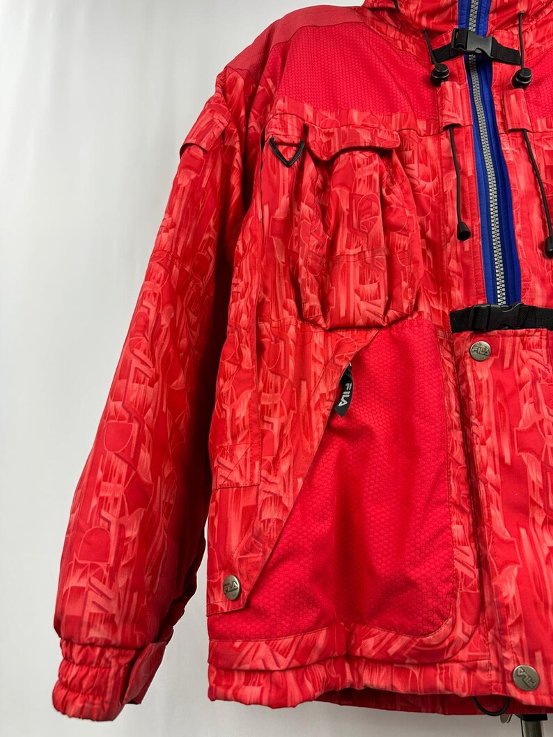 Puede incluir: Chaqueta de esqu&iacute; Fila roja con un patr&oacute;n geom&eacute;trico. La chaqueta presenta una cremallera azul, detalles en negro y m&uacute;ltiples bolsillos. Tiene un cuello alto y cierre de bot&oacute;n.