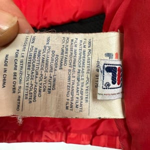 Puede incluir: Primer plano de una etiqueta de una chaqueta roja. La etiqueta tiene el logotipo de la marca y texto en varios idiomas, incluyendo ingl&eacute;s, italiano y alem&aacute;n. La etiqueta indica los materiales de la chaqueta y su origen.