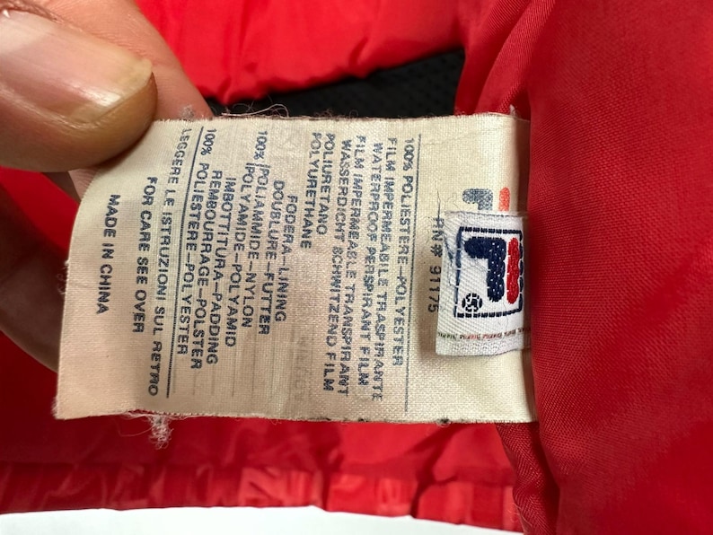 Puede incluir: Primer plano de una etiqueta de una chaqueta roja. La etiqueta tiene el logotipo de la marca y texto en varios idiomas, incluyendo ingl&eacute;s, italiano y alem&aacute;n. La etiqueta indica los materiales de la chaqueta y su origen.