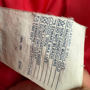 Puede incluir: Primer plano de una etiqueta blanca de cuidado de la ropa con texto y s&iacute;mbolos negros. La etiqueta incluye instrucciones de lavado, como lavar a m&aacute;quina en fr&iacute;o, no usar lej&iacute;a y planchar a baja temperatura. La etiqueta est&aacute; sobre una tela roja.