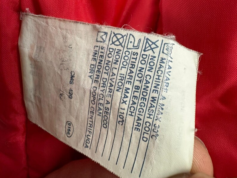 Puede incluir: Primer plano de una etiqueta blanca de cuidado de la ropa con texto y s&iacute;mbolos negros. La etiqueta incluye instrucciones de lavado, como lavar a m&aacute;quina en fr&iacute;o, no usar lej&iacute;a y planchar a baja temperatura. La etiqueta est&aacute; sobre una tela roja.