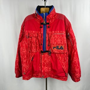 Puede incluir: Chaqueta de esqu&iacute; roja vintage con un patr&oacute;n texturizado. La chaqueta presenta una cremallera, cuello y detalles en azul, con el logo "FILA" en el pecho. Tiene m&uacute;ltiples bolsillos y un cuello alto, adecuada para el fr&iacute;o.