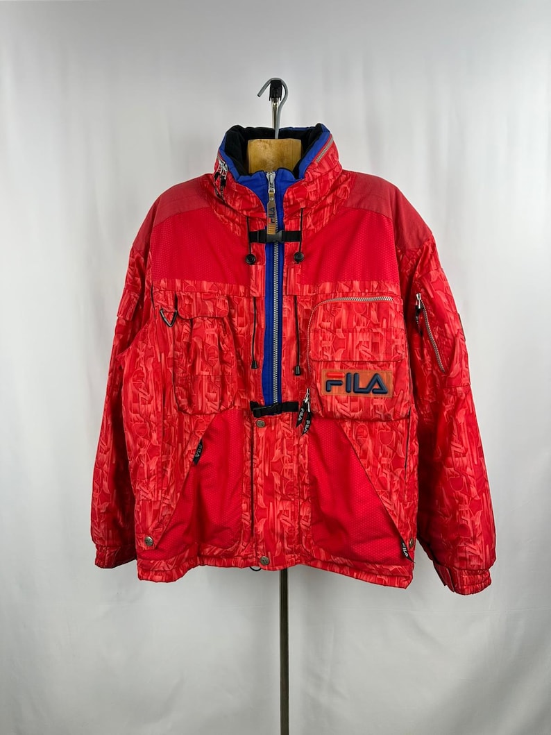 Puede incluir: Chaqueta de esqu&iacute; roja vintage con un patr&oacute;n texturizado. La chaqueta presenta una cremallera, cuello y detalles en azul, con el logo "FILA" en el pecho. Tiene m&uacute;ltiples bolsillos y un cuello alto, adecuada para el fr&iacute;o.