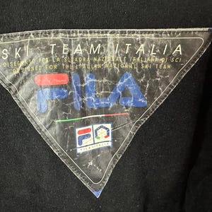 Puede incluir: Primer plano de un parche triangular en blanco y negro con el texto "SKI TEAM ITALIA" y "FILA" en azul y rojo. El parche est&aacute; dise&ntilde;ado para el equipo nacional italiano de esqu&iacute;.
