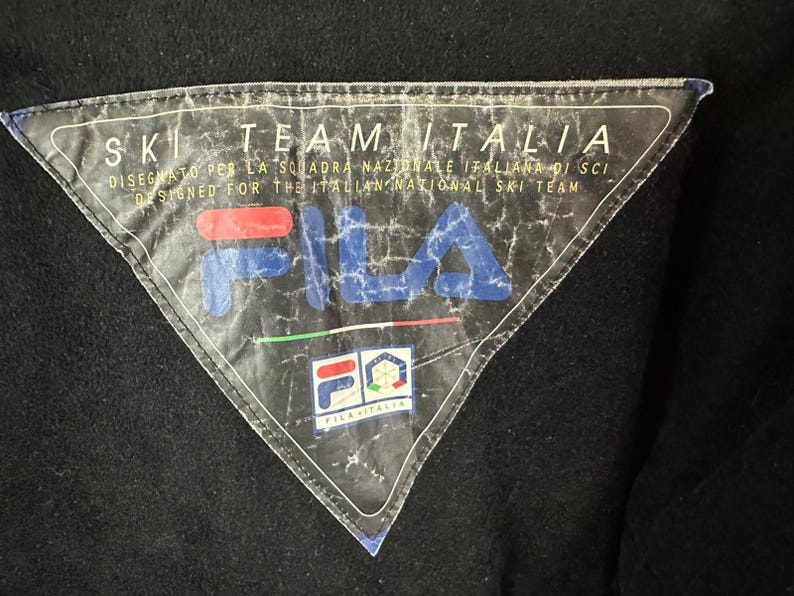 Puede incluir: Primer plano de un parche triangular en blanco y negro con el texto "SKI TEAM ITALIA" y "FILA" en azul y rojo. El parche est&aacute; dise&ntilde;ado para el equipo nacional italiano de esqu&iacute;.