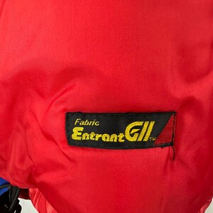 Puede incluir: Primer plano de una tela roja brillante. Una etiqueta rectangular negra con texto amarillo dice "Fabric Entrant GII". La etiqueta tiene un tri&aacute;ngulo rojo y blanco en el lado derecho. La tela parece ser parte de una prenda.