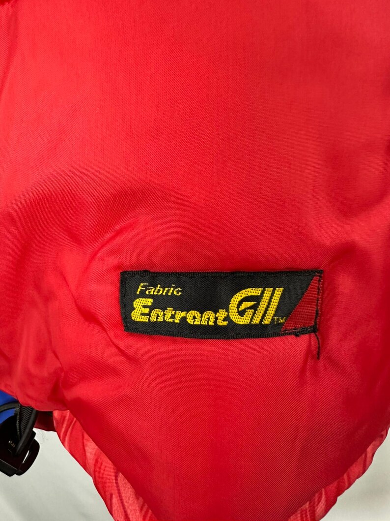 Puede incluir: Primer plano de una tela roja brillante. Una etiqueta rectangular negra con texto amarillo dice "Fabric Entrant GII". La etiqueta tiene un tri&aacute;ngulo rojo y blanco en el lado derecho. La tela parece ser parte de una prenda.