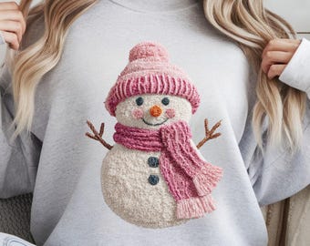 Puede incluir: Una sudadera gris claro con un diseño de muñeco de nieve. El muñeco de nieve tiene un gorro de punto rosa, una bufanda y mejillas rosadas. Tiene brazos de ramitas y una nariz de zanahoria. El diseño está centrado en el pecho.