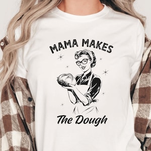 May include: White t-shirt with a black and white graphic of a woman holding bread, wearing glasses and an apron. The text "MAMA MAKES The Dough" is above and below the illustration.