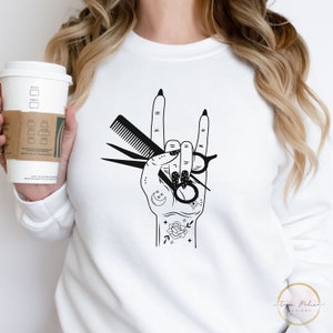 Peut inclure: Un sweat-shirt blanc avec un motif noir d'une main tenant des ciseaux et un peigne. La main fait le geste rock and roll. La main est d&eacute;cor&eacute;e d'un croissant de lune, d'une &eacute;toile et d'une rose.
