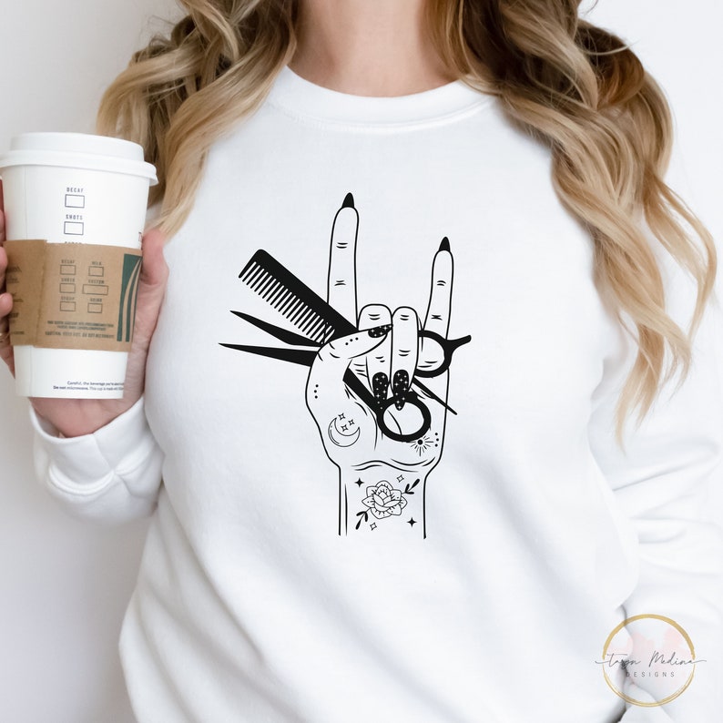 Peut inclure: Un sweat-shirt blanc avec un motif noir d'une main tenant des ciseaux et un peigne. La main fait le geste rock and roll. La main est d&eacute;cor&eacute;e d'un croissant de lune, d'une &eacute;toile et d'une rose.