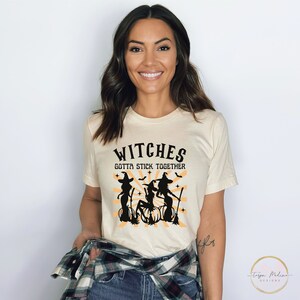 Puede incluir: Una camiseta de color crema con un gr&aacute;fico negro de tres brujas volando en escobas con el texto "Witches Gotta Stick Together".