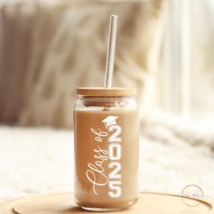 May include: A clear glass tumbler with a brown lid and a straw. The tumbler has a white design that says "Class of 2025" with a graduation cap above the 2.