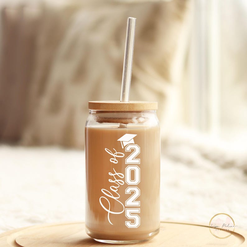 May include: A clear glass tumbler with a brown lid and a straw. The tumbler has a white design that says "Class of 2025" with a graduation cap above the 2.