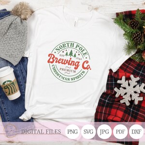 May include: A white t-shirt with a green and red circular design that says "North Pole Brewing Co. Est. 1832 Premium Christmas Spirits".