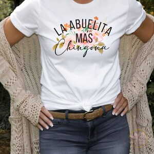May include: White t-shirt with the text "La Abuelita Mas Chingona" in black lettering with a floral design.