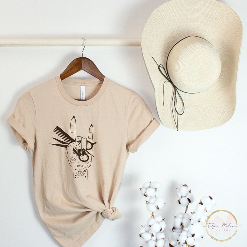 Peut inclure: Un t-shirt beige avec un motif noir d'une main tenant des ciseaux et un peigne dans un geste de rock and roll.