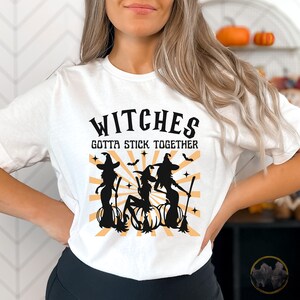 Puede incluir: Camiseta blanca con un gr&aacute;fico negro de tres brujas en escobas con el texto "Witches Gotta Stick Together".