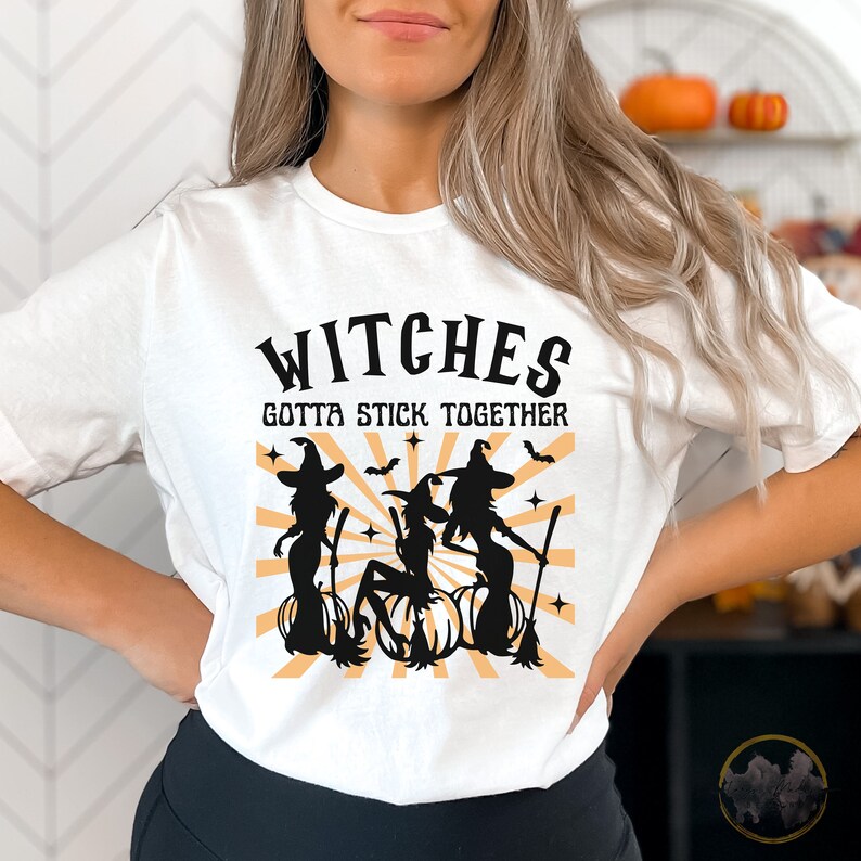 Puede incluir: Camiseta blanca con un gr&aacute;fico negro de tres brujas en escobas con el texto "Witches Gotta Stick Together".