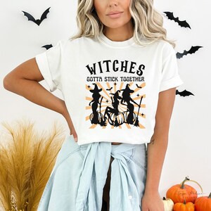 Puede incluir: Camiseta blanca con un gr&aacute;fico negro de tres brujas en escobas con el texto "Witches Gotta Stick Together".