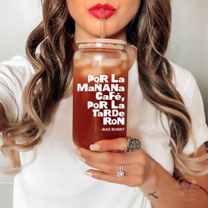 May include: A clear glass tumbler with a brown beverage inside and a white straw. The tumbler has the text "POR La MaNaNa CaF&eacute;, PoR La TaRdE RoN - BAD BUNNY" printed on it.