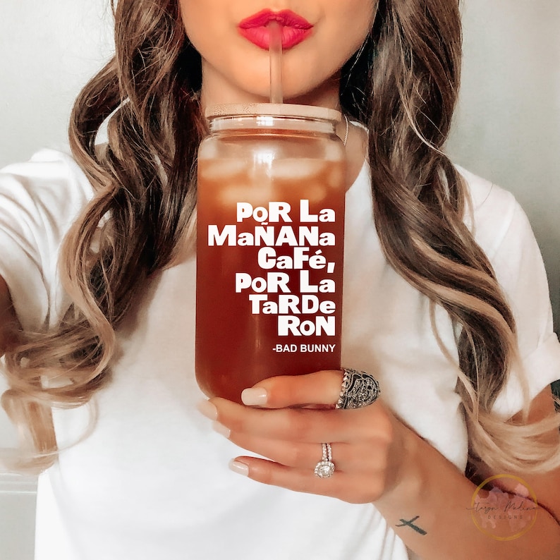 May include: A clear glass tumbler with a brown beverage inside and a white straw. The tumbler has the text "POR La MaNaNa CaF&eacute;, PoR La TaRdE RoN - BAD BUNNY" printed on it.
