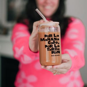 May include: A clear glass tumbler with a bamboo lid and a black straw. The tumbler is filled with a brown liquid and has the text "Por La Ma&ntilde;ana Caf&eacute;, Por La Tarde Ron -Bad Bunny" printed on the side.