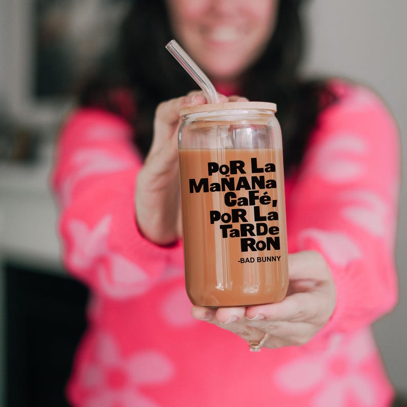 May include: A clear glass tumbler with a bamboo lid and a black straw. The tumbler is filled with a brown liquid and has the text "Por La Ma&ntilde;ana Caf&eacute;, Por La Tarde Ron -Bad Bunny" printed on the side.