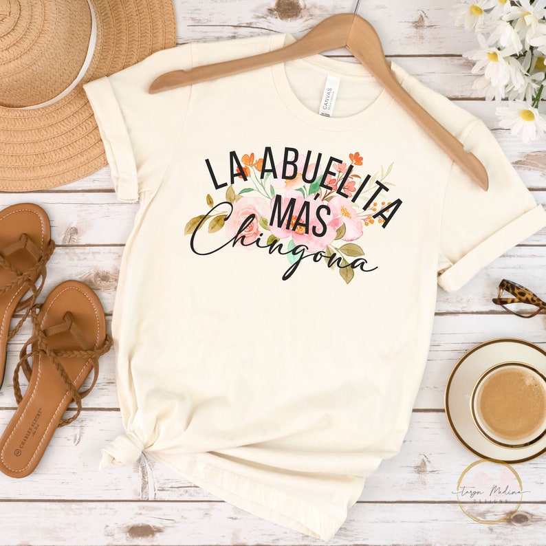 May include: A white t-shirt with a floral design and the text "La Abuelita Mas Chingona" in black.