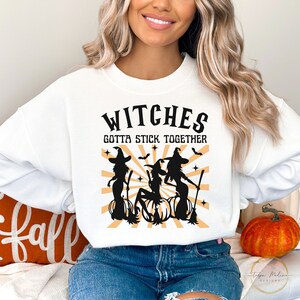 Puede incluir: Sudadera blanca con un gr&aacute;fico negro de tres brujas con escobas y calabazas. El texto "Witches Gotta Stick Together" est&aacute; impreso en negro sobre el gr&aacute;fico.