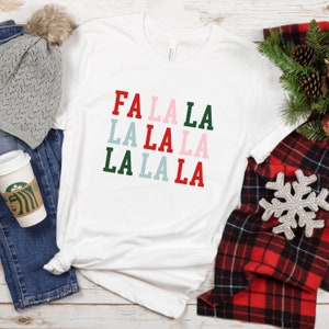 Può includere: Maglietta bianca con il testo "FA LA LA LA LA LA LA LA LA" in rosso, verde, rosa e blu.