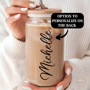 May include: A clear glass reusable coffee cup with a brown lid and a straw. The cup is filled with a brown liquid and has the name "Michelle" written on the side. The text "OPTION TO PERSONALIZE ON THE BACK" is written on the image.