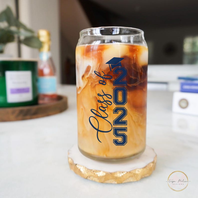 May include: A clear glass can with a blue and white "Class of 2022" design filled with iced coffee. The can is sitting on a white and gold coaster.