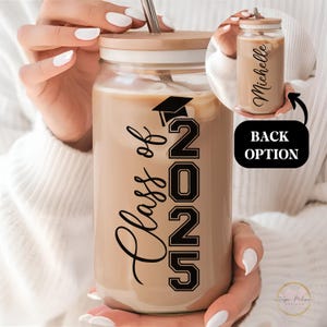 May include: A clear glass mason jar with a wooden lid and a straw. The jar is filled with a brown liquid and has a black vinyl decal that says "Class of 2025" with a graduation cap above the 2. The back of the jar has a white vinyl decal with the name "Michelle".