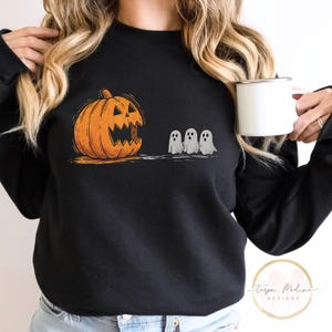 Puede incluir: Sudadera negra con un gráfico de Halloween. El diseño incluye una calabaza naranja con una cara tallada y tres pequeños fantasmas blancos. La sudadera es de cuello redondo.