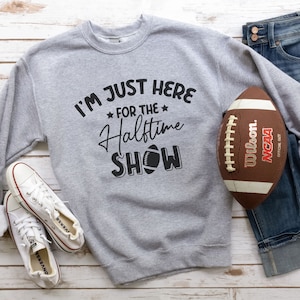 Op de afbeelding: Een grijs sweatshirt met de tekst "I'm just here for the halftime show" en een voetbalafbeelding. Op de achtergrond zijn een bruine voetbal en een blauwe jeans te zien.