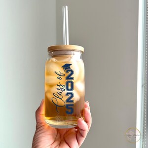 May include: A clear glass mason jar with a wooden lid and a straw. The jar is filled with ice and a brown liquid. The jar has a blue and white decal that says "Class of 2025" with a graduation cap.