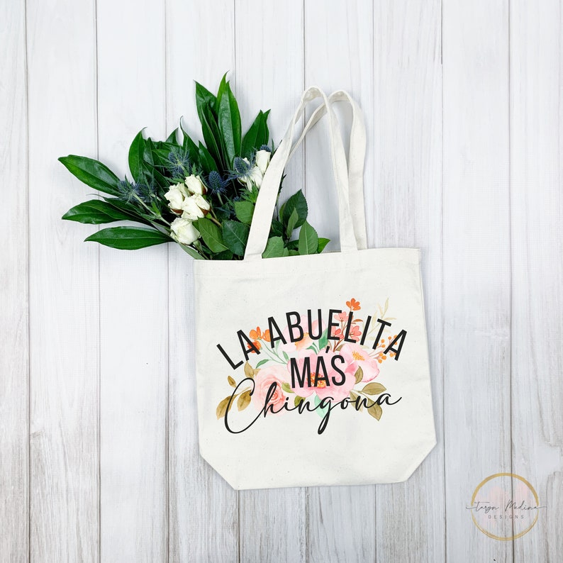 May include: A white canvas tote bag with a floral design and the text "La Abuelita Mas Chingona" in black.