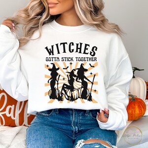 Puede incluir: Sudadera blanca con el texto "Witches Gotta Stick Together" en negro. El dise&ntilde;o presenta tres brujas en siluetas negras con escobas y calabazas sobre un fondo de rayos naranja y amarillo.