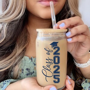 May include: A clear glass tumbler with a bamboo lid and a blue and white "Class of 2023" design. The tumbler is filled with a brown beverage and a clear drinking straw.