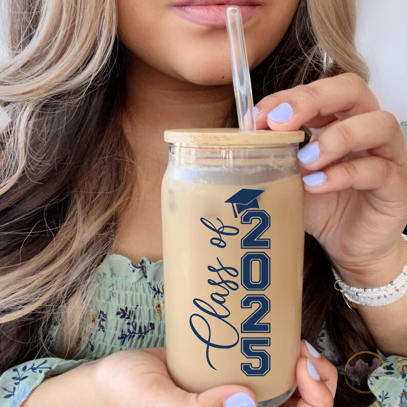 May include: A clear glass tumbler with a bamboo lid and a blue and white "Class of 2023" design. The tumbler is filled with a brown beverage and a clear drinking straw.